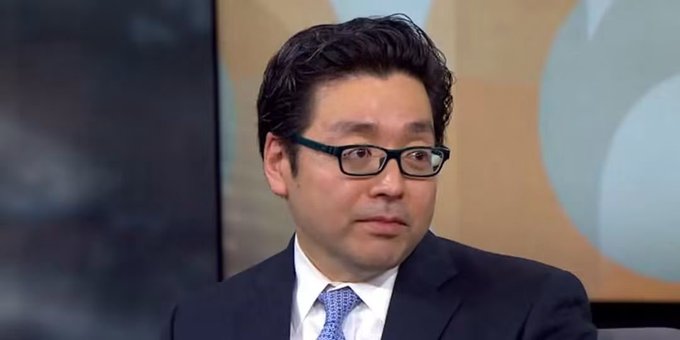 A man with dark hair and glasses wearing a dark suit and blue patterned tie appears to be speaking during a television interview.