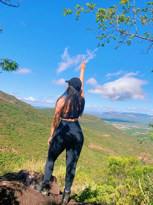 The image features a woman standing on top of a hill, wearing black clothing and holding her arms up in the air while looking out over the landscape below. The sky above them appears blue with some clouds scattered across it. In the background, there are mountains visible, adding depth to the scene. The post seems to be promoting an event or gathering related to web3 technology, as indicated by the text "good morning @base good morning @Polymarket Creator Active".