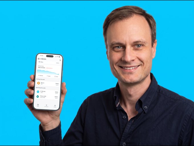 A smiling man in a dark shirt holds up a smartphone displaying the AQUA app interface against a bright blue background, showcasing account balances and transaction history. The image visually reinforces the post's message by depicting the creator of Bitcoin futures personally using the AQUA platform.
