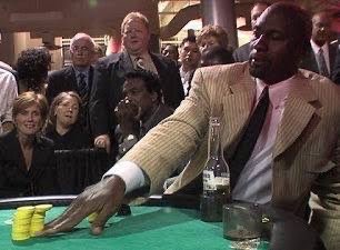 A low-resolution, grainy video still showing a man in a tan suit at what appears to be a craps or casino table, reaching down to handle yellow chips.