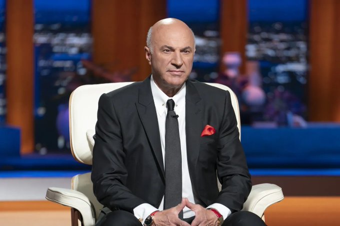 Kevin O'Leary from the TV show Shark Tank, wearing a dark suit with a red pocket square, sitting in a white chair on the show's set.