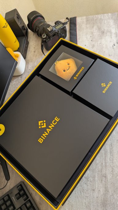 A unboxing photo showing a black 'BINANCE' branded box on a desk alongside a camera with a red ring lens and other tech accessories.