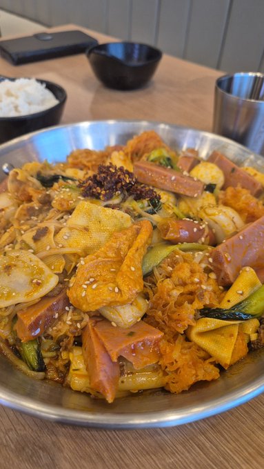 A large metal bowl filled with a spicy Korean stir-fried dish (appears to be rabokki or tteokbokki) containing rice cakes, spam, noodles, vegetables, and chili flakes, served with a side of rice.