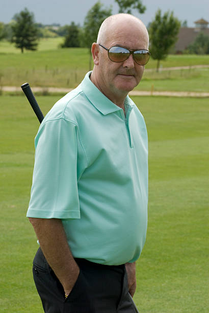 A photograph of an older, bald man wearing sunglasses, a teal polo shirt, and dark trousers, standing on a golf course with a golf club slung over his shoulder.