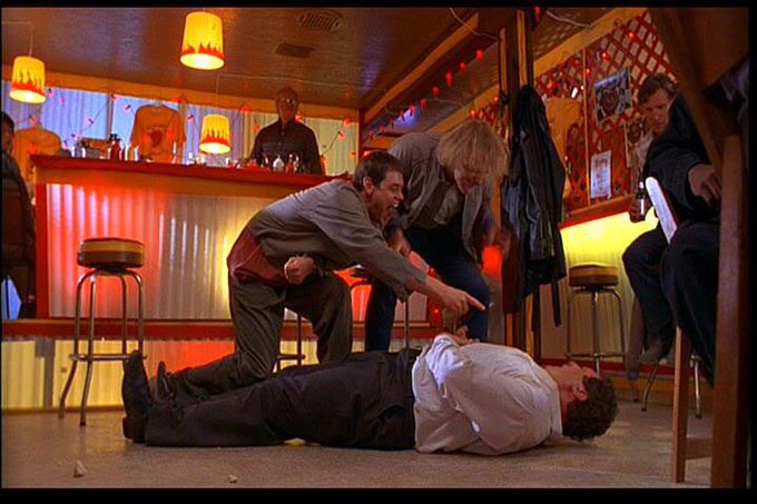 In a dimly lit, neon-accented bar, a man in a white shirt lies sprawled on the floor while another man in a gray jacket points down at him accusingly, with onlookers watching the tense scene unfold. The image humorously mirrors the post's story of a reckless trader repeatedly overleveraging only to be liquidated each time, capturing the moment of realization and frustration.