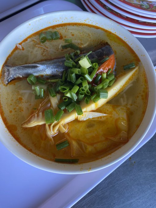 A steaming bowl of Vietnamese fish soup, topped with fresh green onions and featuring a whole fish and bamboo shoots in a rich orange broth, is served on a white plate beside stacked red-rimmed bowls, capturing a hearty breakfast meant to recharge after a long night out.