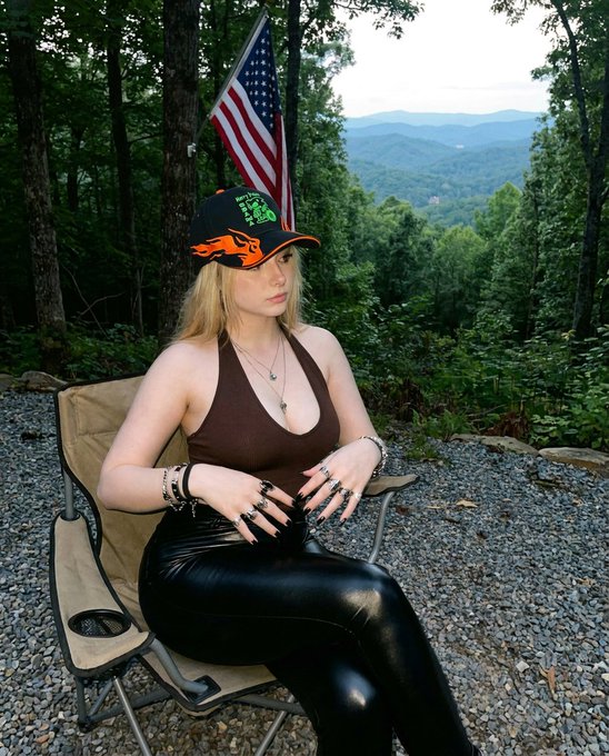 A blonde woman wearing a black and orange cap, brown halter top, and black leather pants sits in a camping chair outdoors with an American flag and a scenic mountain valley view in the background.