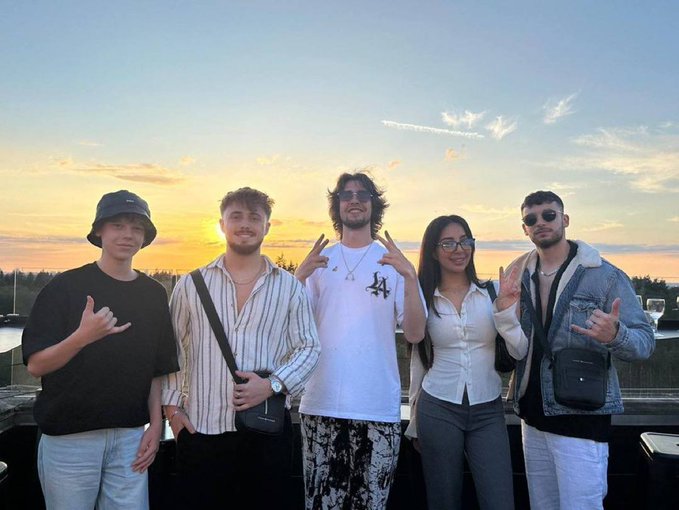 Five friends pose together at sunset with a vibrant orange sky behind them, flashing hand gestures and smiles on what appears to be a rooftop or elevated outdoor venue. The "GM" (Good Morning) post is a common Web3/crypto social media greeting, and the casual group photo captures the friendly, community-driven spirit of the crypto space.