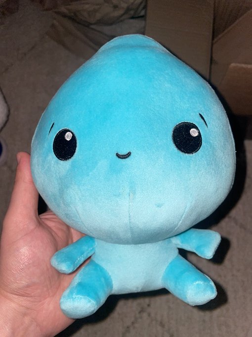 A hand holds a small, light blue plush toy with a teardrop-shaped head, large black eyes, and a small smile against a carpeted background.