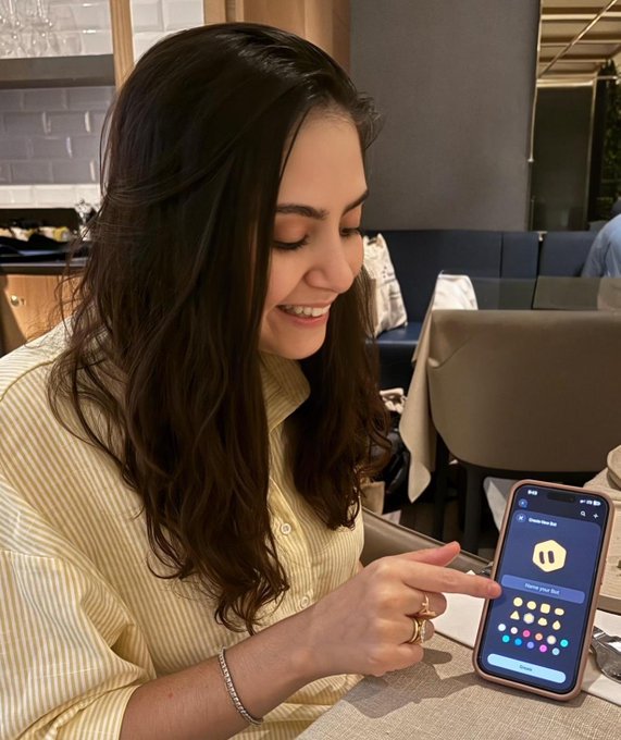 A smiling woman with long dark hair, wearing a yellow striped shirt and silver bracelets, sits in a restaurant while showing her smartphone displaying the "Grok" app onboarding screen with a "Choose Your Bot" prompt. The image captures a candid, intimate date night moment as she engages with the new AI bot feature.