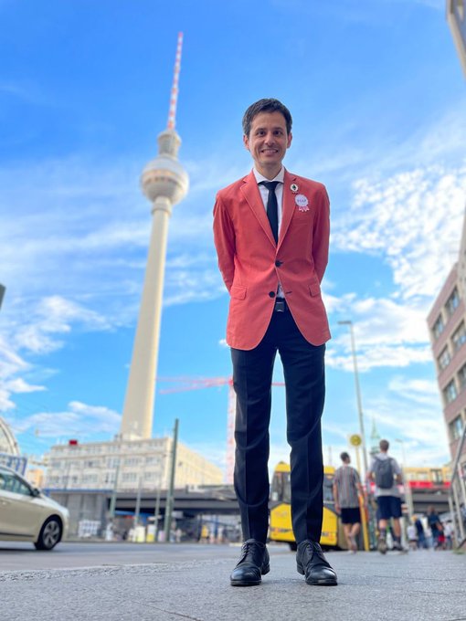 A man in a coral blazer and dark tie stands smiling in front of Berlin's iconic TV Tower (Fernsehturm), with a bright blue sky and construction cranes visible in the background.

The image appears to capture a memorable moment with this POAP-distributing figure at an Ethereum conference in Berlin, as the post nostalgically reflects on the end of an era of receiving physical POAP badges at such events.