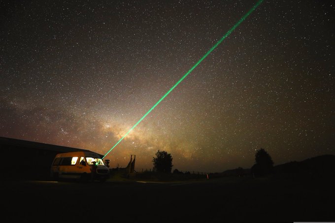 The image features a van parked under a starry night sky with a green laser beam coming from its rear window. The scene also includes a person standing nearby, possibly observing or participating in an activity related to the laser beam. The focus of the image is on the unique and intriguing nature of the laser beam, which stands out against the backdrop of the dark sky.