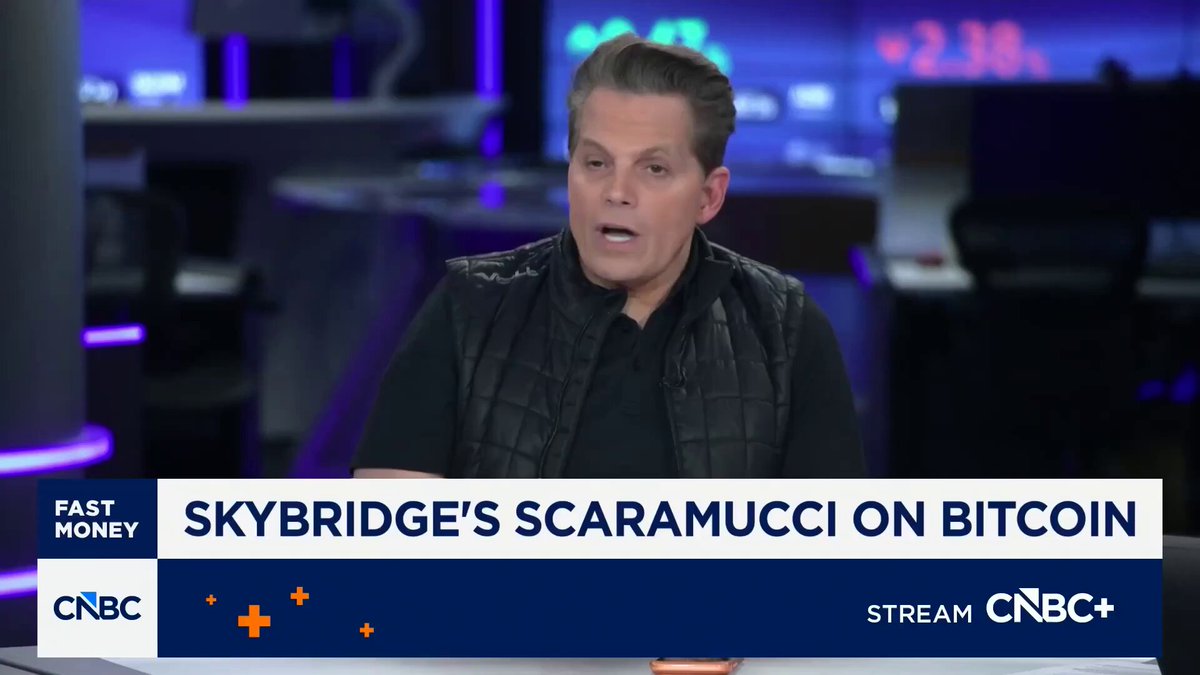 The image features a man wearing a black vest, sitting at a desk with his hands on his knees while talking on the phone. The background of the image shows a TV screen displaying a news segment about Bitcoin. The text on the image reads "Anthony Scaramucci predicts Bitcoin rally will begin late Q4 202 into early 2027". This suggests that the man might be discussing the potential rise in Bitcoin prices during the fourth quarter and early twenty-first century, as indicated by the news segment.