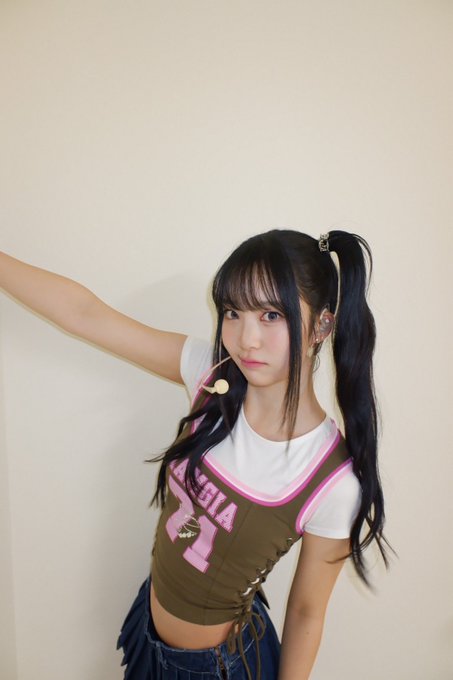 A young woman with long dark twin-tail pigtails wears a white t-shirt and a brown crop top with pink trim, posing with one arm extended against a plain wall.