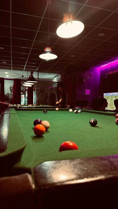 A dimly lit billiards room featuring a green felt pool table with scattered billiard balls, illuminated by hanging lamps under a dark ceiling.