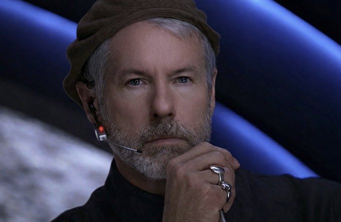 The image features a man with gray hair wearing a brown hat and holding a microphone close to his face. The background consists of blue stripes or lights that create an interesting visual effect, making the scene appear more dynamic and engaging. There are also some words visible in the image, which could be related to Bitcoin trading or cryptocurrency news.