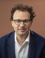 The image shows a man with curly hair and glasses wearing a dark blazer over a white shirt, likely a financial commentator or analyst. His professional appearance aligns with the accompanying post announcing that the S&P 500 ($SPX) has hit his predicted $7,800 target, while warning that new all-time highs could signal an upcoming market trap rather than safety.