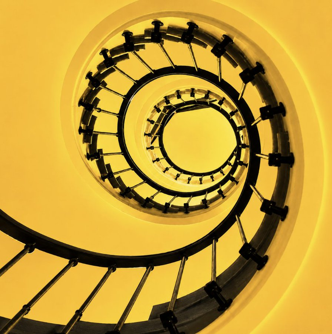 A spiral staircase viewed from above, bathed in warm yellow light, perfectly visualizing the metaphor of progress moving in an upward spiral. Its continuous, winding steps echo the idea that growth and development advance in waves, reinforcing the message of perseverance and forward momentum.