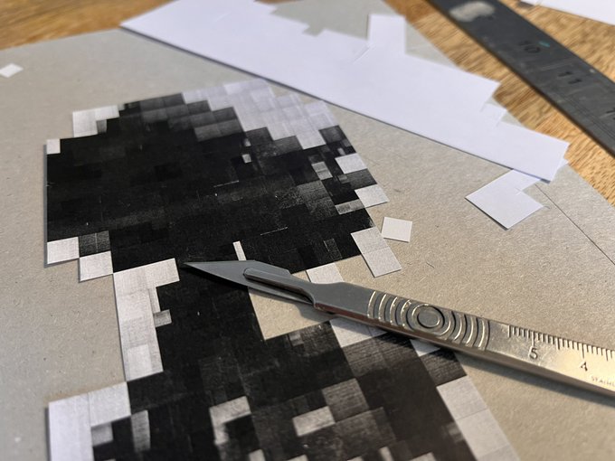 A close-up of a craft cutting process showing a metal precision knife and ruler cutting pixelated black and white artwork on a craft mat.