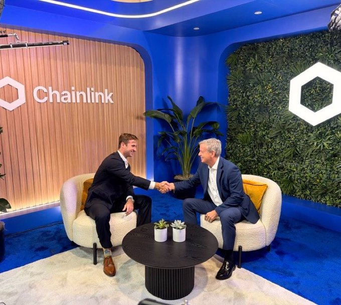 Two men in suits shake hands in a branded Chainlink studio space featuring blue walls, a 'Chainlink' logo display, and a green plant wall with a hexagonal logo.