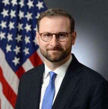 A portrait of a bearded man in a suit and glasses standing before an American flag, identified by the post as White House Director Patrick Witt amid reports about potential Bitcoin acquisitions for the U.S. Strategic Reserve.
