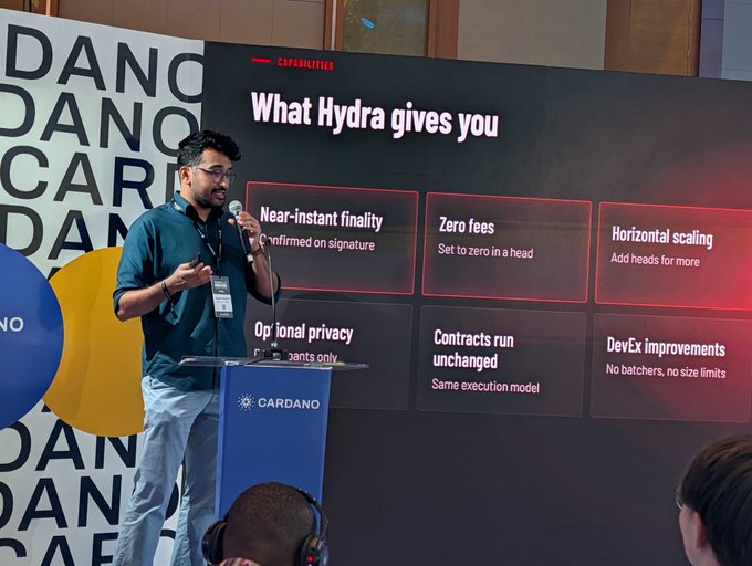 A speaker presents at a Cardano event beside a slide titled “What Hydra gives you,” listing benefits including “Near-instant finality,” “Zero fees,” “Horizontal scaling,” “Optional privacy,” “Contracts run unchanged,” and “DevEx improvements.” The scene aligns with the post’s message about discussing Hydra’s fast off-chain transactions and final settlement on Cardano at Token2049.