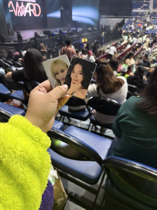 A concertgoer wearing a fuzzy yellow sweater holds up two photocards of MAMAMOO members in an arena, with "AWARD" illuminated on a stage in the background. The image reflects the post's reference to MAMAMOO members SOLAR, WHEE BYUL, and HWASA, likely taken at the 2024 MAMAMOO fan gathering or related awards event.