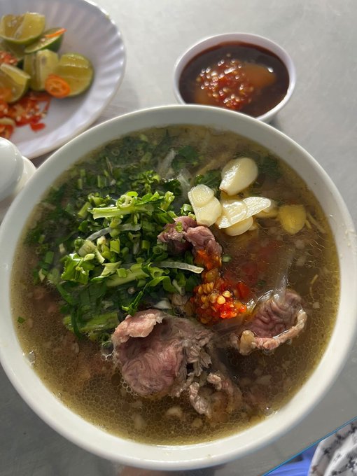 A bowl of Vietnamese-style beef pho topped with herbs, onions, and chili, accompanied by a plate of lime wedges and a side of chili sauce.