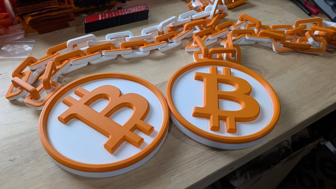 3D-printed orange and white Bitcoin coin models connected by chains, displayed on a wooden workbench.