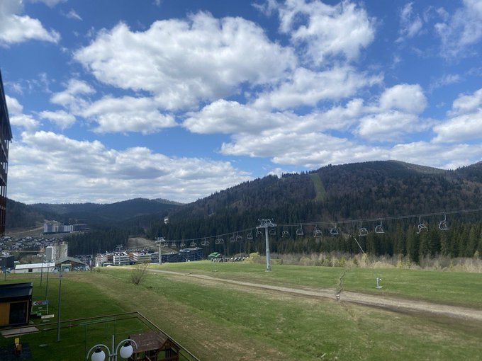 A mountain resort landscape view showing a ski lift with empty chairs running above a green grassy slope, with a town nestled in the valley and forested mountains under a partly cloudy sky.