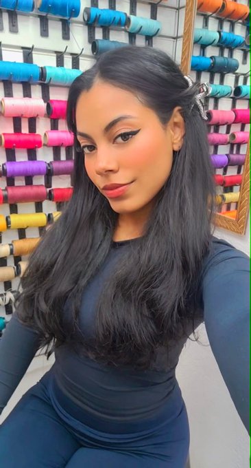 A woman with long dark hair poses for a selfie in what appears to be a fabric or thread store, with colorful spools of thread visible on the wall behind her.