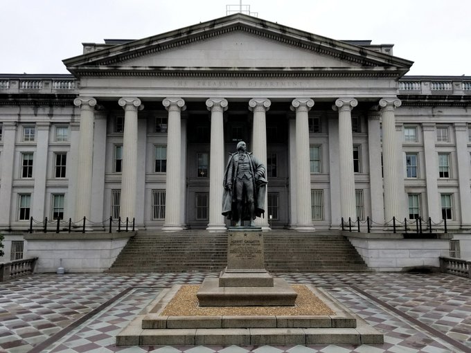 The image shows the U.S. Treasury Department building in Washington, D.C., featuring its iconic neoclassical façade with tall columns and a bronze statue of Alexander Hamilton on a pedestal in the foreground, fittingly linking to the Treasury's recent proposal defining stablecoin issuer regulations under the GENIUS Act.
