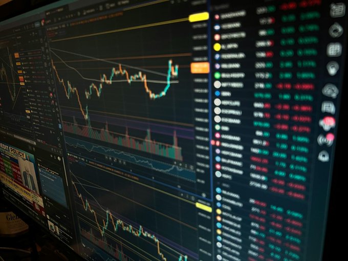A computer monitor displaying financial trading charts with candlestick patterns and a watchlist of various stock/crypto tickers with green and red price changes.