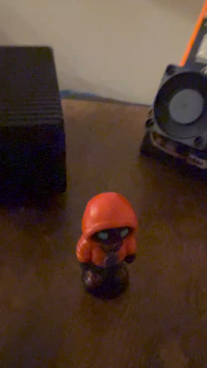 In this image, there's a small figurine of someone wearing an orange hoodie, standing alone on top of a brown surface with white text. The image is taken from a low angle, giving the figurine an imposing presence. There are also two other items in the image - a speaker and a book, which seem unrelated to the main subject of the figurine.
