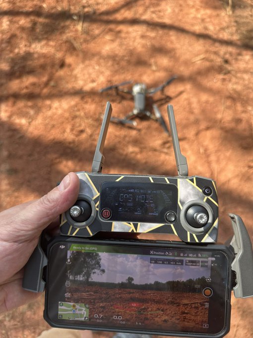 A person holds a drone controller with an attached smartphone showing a GPS mode '0.7' and a live feed of red dirt terrain, while a gray drone hovers nearby on dry ground.