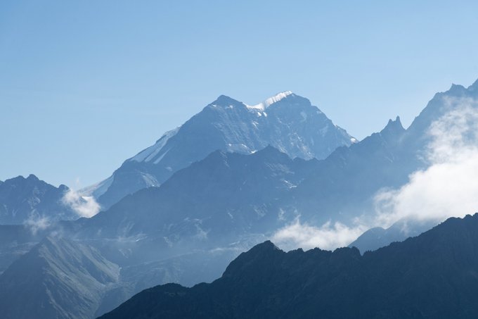 A scenic photograph of majestic, snow-capped mountain peaks partially obscured by wispy clouds against a clear blue sky.