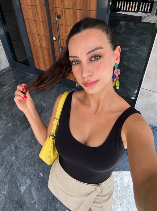 A selfie of a woman wearing a black sleeveless top, beige skirt, colorful beaded earrings, and carrying a yellow shoulder bag while holding a strand of her hair.