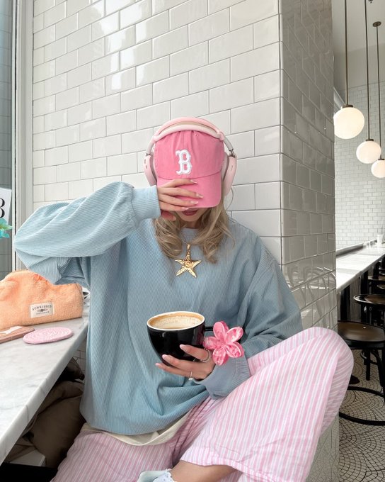 A lifestyle photo of a person in a pastel aesthetic outfit wearing a pink baseball cap with a 'B' logo, blue sweatshirt, and pink striped pants, holding a coffee cup while covering their face with their hand in a cafe setting.