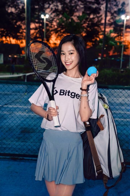 The image features a young woman holding a tennis racket and ball while standing next to a fence or chain link fence, possibly at a tennis court. She appears to be posing for a photo with her tennis gear. The caption of the image reads "Happy Monday, who'S up for a matching #Bitget VIP limited-edition tennis set? Playing tennis is just like trading: the serve is your entry timing, an ACE is a perfect bottom-fishing opportunity, and a double fault is the cost of FOMO-chasing highs". The image seems to be related to the post about trading in the stock market.