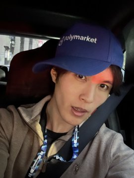 A selfie of a person wearing a blue "Polymarket" cap and a lanyard, taken inside a vehicle.