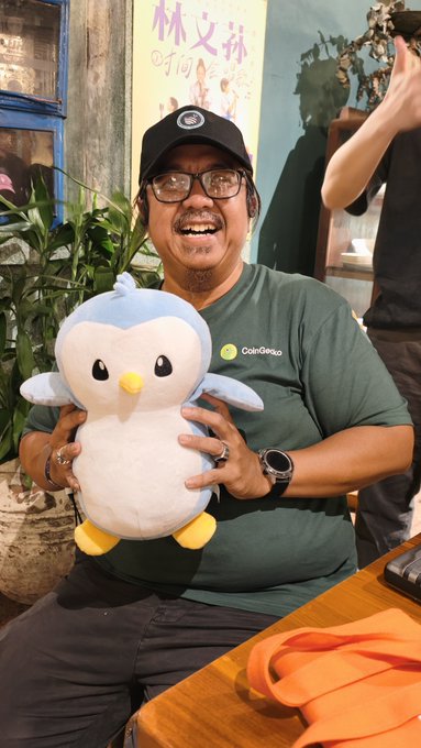 A smiling man wearing a green 'CafeGecko' t-shirt, black cap, and glasses holds up a blue and white penguin plushie.