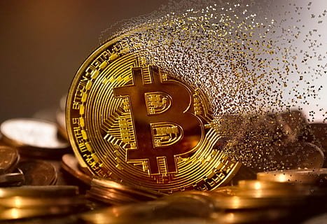 A gold Bitcoin coin with particles dispersing from it, surrounded by other coins.