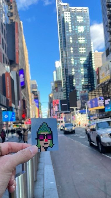 A hand holds up a small pixel-art sticker of a green-haired character with sunglasses against the backdrop of a bustling city street with tall skyscrapers and billboards.