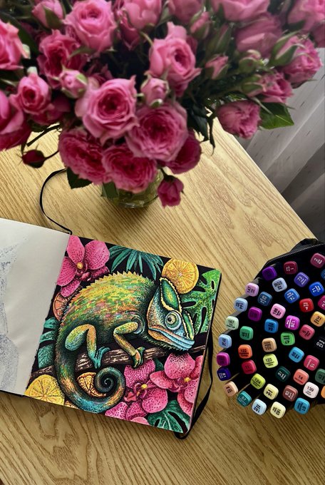 This image is related to the Instagram post it is a hashtag for #Tezostuesday. The photo shows an open notebook with a detailed illustration of a lizard in a tropical scene. Surrounding the notebook are art supplies and a bouquet of roses.