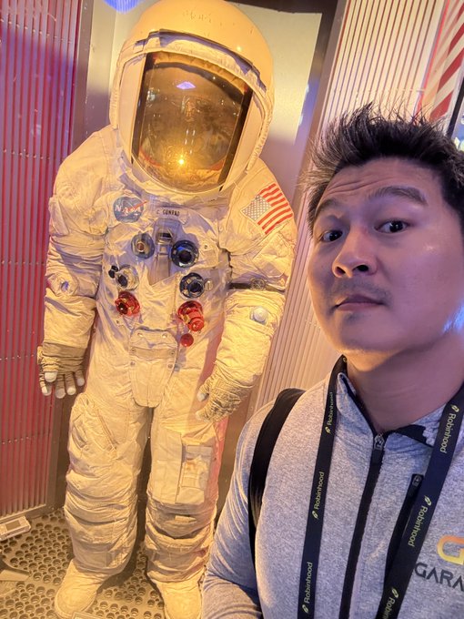 A man wearing a "TeamProved" lanyard poses for a selfie next to a full-size NASA astronaut spacesuit display, with the gold-visored helmet and American flag patch clearly visible, fitting the "going to the moon" theme of his post.