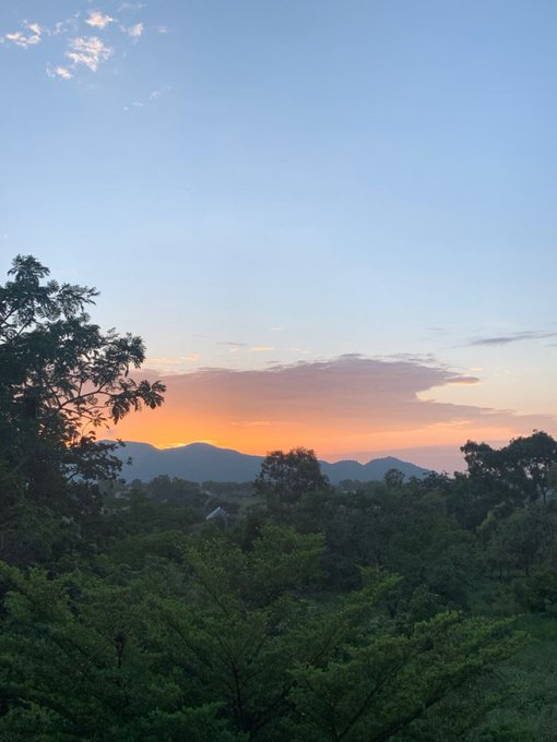 A serene landscape view at sunset, featuring lush green trees in the foreground and silhouetted mountains against a gradient blue and orange sky.