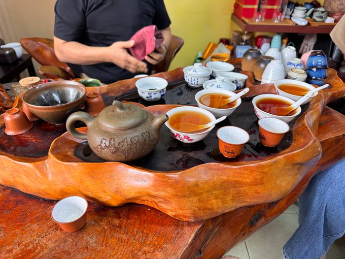 A person wipes a small bowl with a pink cloth at a traditional Chinese tea ceremony, surrounded by a large wooden tea tray laden with a clay teapot, various small teacups filled with amber tea, and ceramic tea accessories.