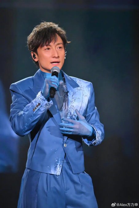 A male performer in an elegant blue satin suit and matching gloves sings into a microphone on stage, with a watermark "@Alex力申" visible in the corner. While the accompanying post discusses Jan Lamb's NFT investment losses, the image itself only shows a singer mid-performance and does not depict Jan Lamb or any visual reference to cryptocurrency or NFTs.
