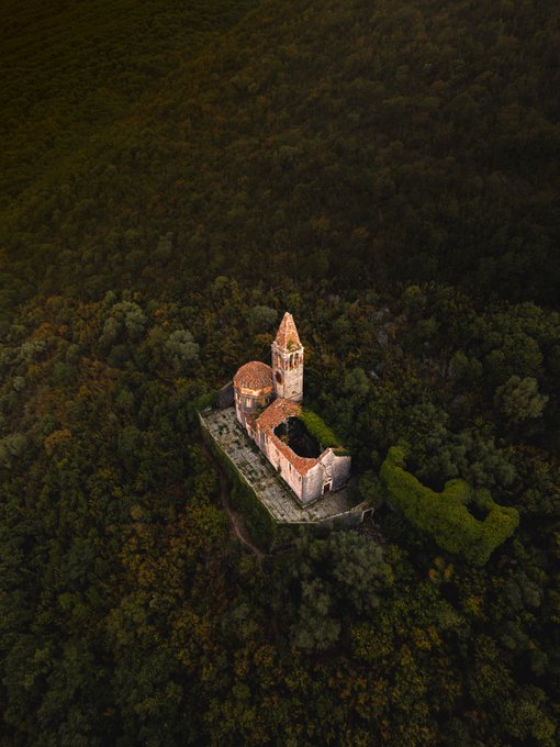 An aerial view of an old stone church with a tall tower and red-tiled roof, surrounded by dense forest.
