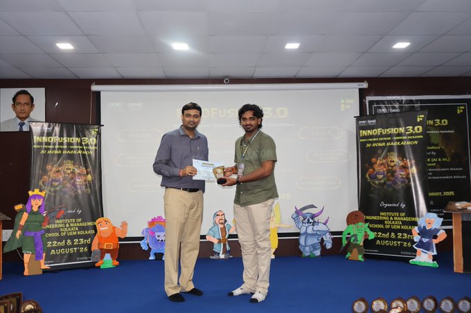 Two men stand on stage at the InnoFusion 3.0 event at UCSI University Kuala Lumpur, exchanging an award, with cartoon character cutouts lining the front of the stage.